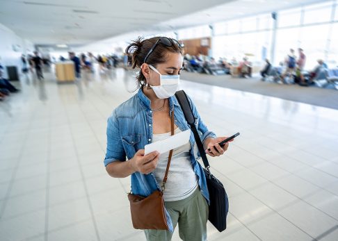 Woman looking at phone for travel cancellations at airport while wearing face mask
