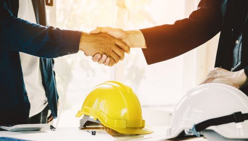 Two men shaking hands over a construction deal with a hard hat in foreground